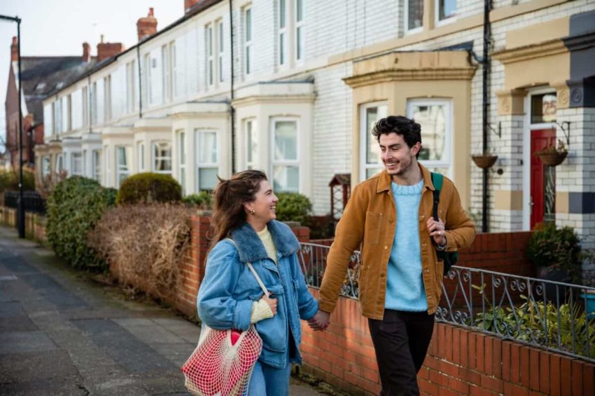Front view of a young couple walking down terraced Street in Whitley Bay in the north-east of England they are heading into the town centre and deciding which shops to go to they are also holding hands and carrying bags over their shoulders.