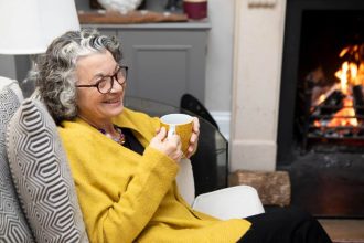 A mature adult sitting by a fireplace in a living room at home. She is wearing a yellow cardigan and spectacles.